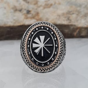 May include: Oval-shaped silver ring with a black center featuring a white geometric design. The ring has a gold-colored inner band and is surrounded by a detailed silver border with stars and a leaf pattern.