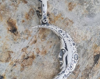 Puede incluir: Un collar con un colgante de media luna de plata, con una superficie texturizada y un diseño de rostro. El colgante está unido a una cadena de plata.