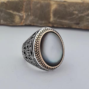May include: A silver ring with an oval-shaped mother-of-pearl stone set in a gold-toned bezel. The ring features intricate silver filigree designs.