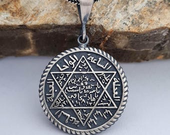 May include: Silver necklace with a round pendant featuring a six-pointed star with Arabic script. The pendant has a rope-like border.