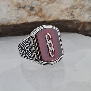 Op de afbeelding: Een zilveren ring met een rechthoekige, bordeauxrode steen. Een zilveren kettingontwerp is gecentreerd op de steen. De ring heeft gedetailleerde patronen aan de zijkanten en een gepolijste afwerking.