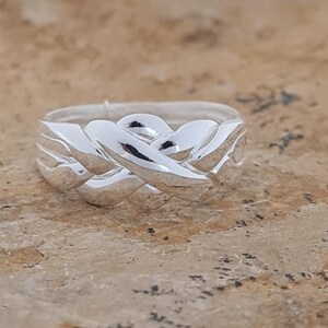 May include: A silver ring with an intricate woven design. The ring's surface reflects light, highlighting the interwoven pattern. The ring appears to be made of a shiny, polished metal, and is a piece of jewelry.
