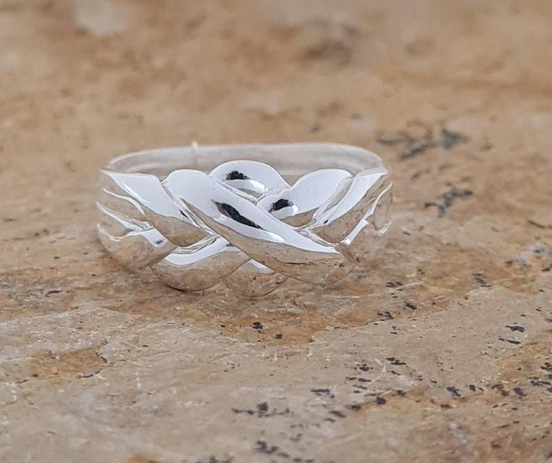 May include: A silver ring with an intricate woven design. The ring's surface reflects light, highlighting the interwoven pattern. The ring appears to be made of a shiny, polished metal, and is a piece of jewelry.