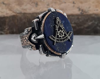 Könnte beinhalten: Silberfarbener Ring mit einem dunkelblauen, ovalen Stein. Der Stein zeigt ein goldfarbenes Freimaurer-Symbol. Der Ring hat aufwendige Details mit goldenen Akzenten. Der Ring wird auf einer reflektierenden Oberfläche präsentiert.