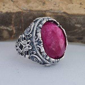 May include: A silver ring with an ornate design featuring a large, oval-shaped, pink gemstone.