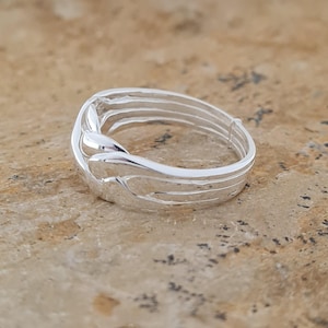 May include: A silver ring with a unique, flowing design. The ring features multiple parallel bands that curve and intertwine, creating an elegant and modern look. The polished metal reflects light, highlighting the intricate details of the ring's construction.