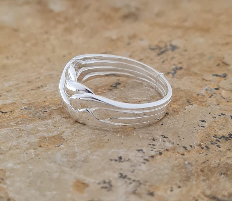 May include: A silver ring with a unique, flowing design. The ring features multiple parallel bands that curve and intertwine, creating an elegant and modern look. The polished metal reflects light, highlighting the intricate details of the ring's construction.
