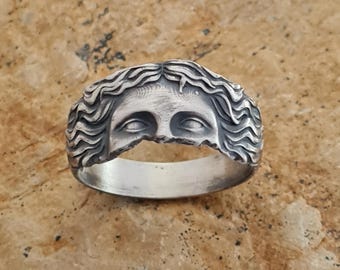Op de afbeelding: Een zilveren ring met een gedetailleerd ontwerp van een gezicht met golvend haar. Het gezicht is in de ringband gegraveerd.