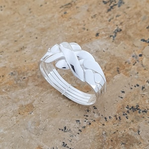 May include: A silver ring with a braided design. The ring features a woven pattern on the top, with multiple bands forming the ring's base. The ring is set against a textured, light brown surface.