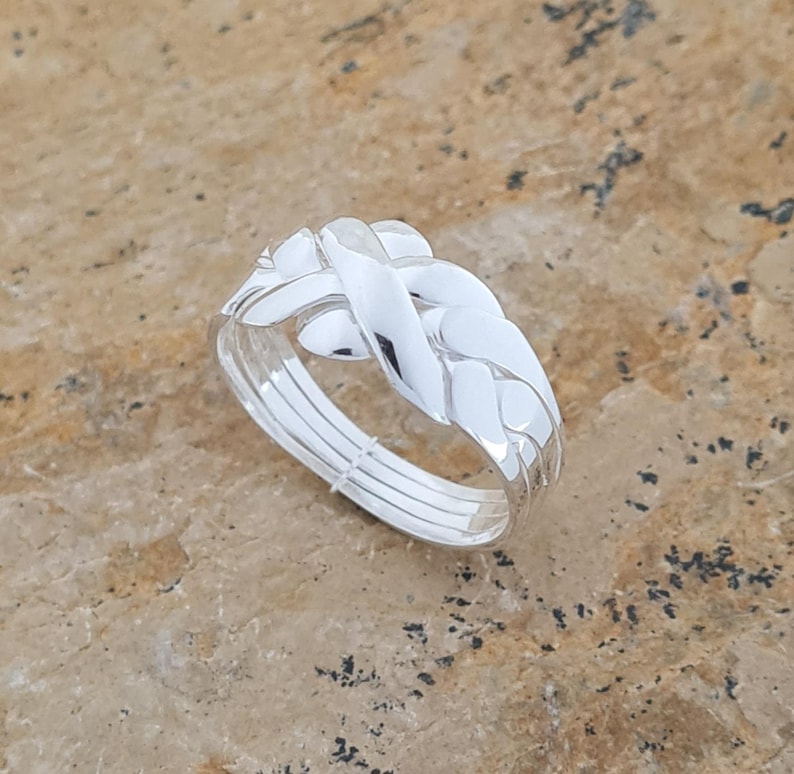 May include: A silver ring with a braided design. The ring features a woven pattern on the top, with multiple bands forming the ring's base. The ring is set against a textured, light brown surface.