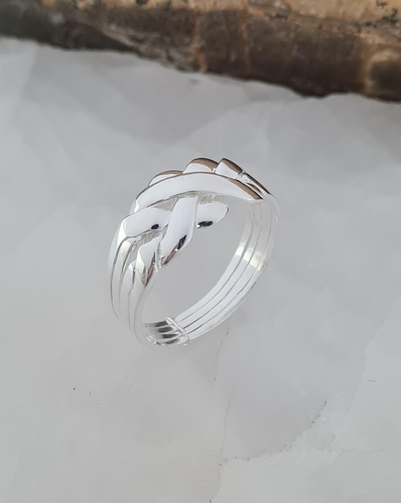 May include: A silver puzzle ring with an interwoven design. The ring features multiple bands and interlocking pieces, creating a complex and intriguing appearance. The polished metal reflects light, highlighting the intricate details of the design.