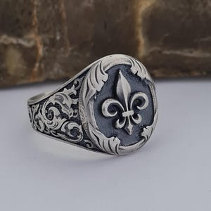 May include: A silver ring with an ornate design featuring a fleur-de-lis in the center. The ring has a black enamel background and a silver band.