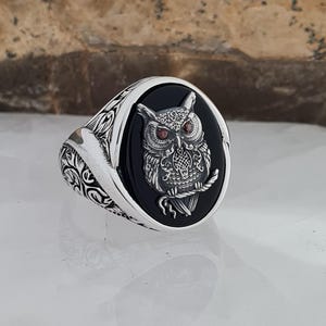 Könnte beinhalten: Silberring mit einem ovalen schwarzen Onyxstein, der ein detailliertes Eulen-Design zeigt. Die Eule hat rote Edelsteinaugen und sitzt auf einem Ast. Das Band des Rings weist ein verziertes, wirbelndes Muster auf.