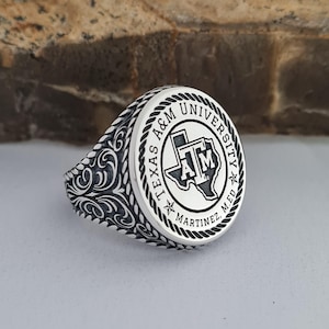 May include: A silver ring featuring the Texas A&M University logo. The ring has a detailed design with the university name and a Texas outline. The band has an ornate, swirling pattern.
