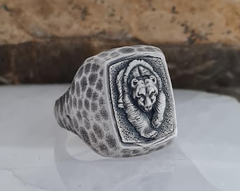 May include: A silver-colored ring with a textured band and a rectangular face. The face features a detailed relief of a bear. The ring has a hammered texture on the band and a polished finish on the bear.
