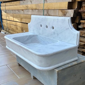 Könnte beinhalten: Ein wandmontiertes Waschbecken aus weißem Marmor mit einem rechteckigen Becken, abgerundeten vorderen Ecken und einer hohen Rückwand. Die Rückwand hat drei kreisförmige Löcher für die Armaturen, und der Stein zeigt eine feine graue Maserung.