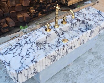 May include: A rectangular white marble sink featuring prominent dark purple veining and a fluted exterior edge. The basin is equipped with a polished gold-tone faucet and two matching handles, along with a gold-tone drain cover centered in the basin.