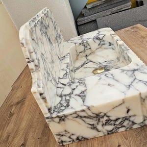 May include: A white marble sink with gray veining. The sink has a rectangular basin and a back splash with a decorative edge. A gold-colored drain is visible in the basin. The sink is made of stone.