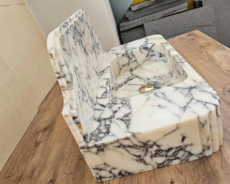 May include: A white marble sink with gray veining. The sink has a rectangular basin and a back splash with a decorative edge. A gold-colored drain is visible in the basin. The sink is made of stone.