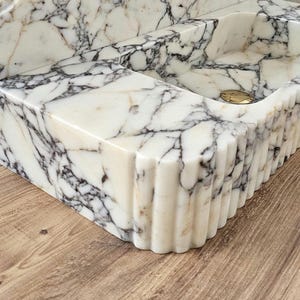 May include: A white marble sink with gray veining. The sink has a rectangular basin and a decorative, fluted edge. A gold-colored drain cover is visible in the basin. The sink is a bathroom fixture.
