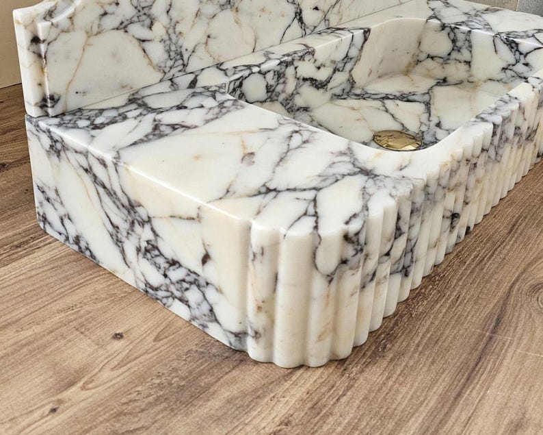 May include: A white marble sink with gray veining. The sink has a rectangular basin and a decorative, fluted edge. A gold-colored drain cover is visible in the basin. The sink is a bathroom fixture.