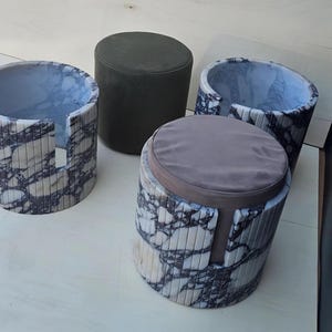 Puede incluir: Cuatro taburetes cilíndricos. Dos están hechos de material similar al mármol con vetas grises y blancas. Uno tiene un cojín gris. El otro es de color verde oscuro liso. Los taburetes tienen un diseño moderno.