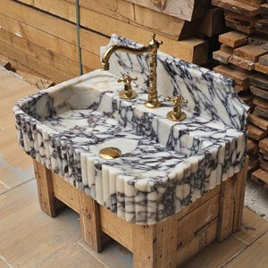 May include: A rectangular marble sink featuring a white and deep purple veined pattern with a fluted exterior design. The sink includes a polished gold-toned faucet and two matching handles, along with a gold-toned drain cover centered in the basin.