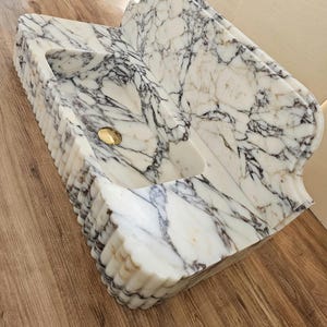 May include: A white marble sink with gray and brown veining. The sink has a rectangular basin with a curved back and a gold-colored drain. The front edge has a fluted design.