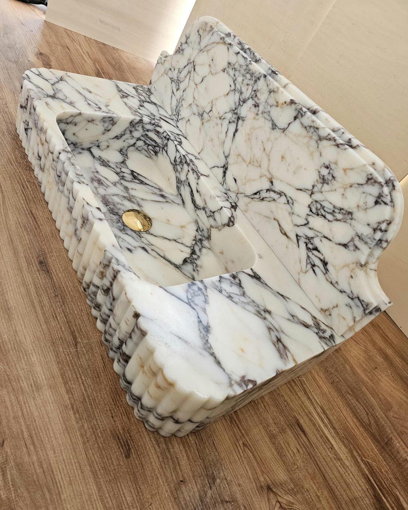 May include: A white marble sink with gray and brown veining. The sink has a rectangular basin with a curved back and a gold-colored drain. The front edge has a fluted design.