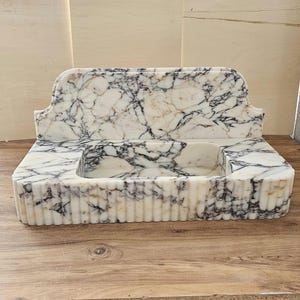 May include: A rectangular marble sink with a white base and black veining. The sink has a built-in backsplash and a basin. The front of the sink has vertical grooves. The sink is on a wooden surface.