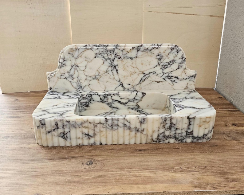 May include: A rectangular marble sink with a white base and black veining. The sink has a built-in backsplash and a basin. The front of the sink has vertical grooves. The sink is on a wooden surface.