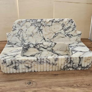 May include: A marble sink with a rectangular basin and a decorative back. The sink is white with black and gray veining. The front of the sink has vertical ribbed detailing.