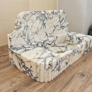 May include: A white marble sink with black veining. The sink has a rectangular basin with a fluted front and a decorative back. The sink is made of stone and has a classic design.