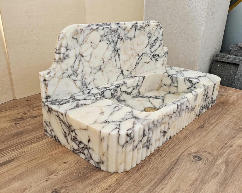 May include: A white marble sink with black veining. The sink has a rectangular basin with a fluted front and a decorative back. The sink is made of stone and has a classic design.