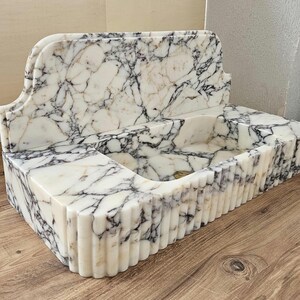 May include: A rectangular marble sink with a white and gray veined pattern. The sink has a curved backsplash and a fluted front. The sink is made of stone and is designed for a bathroom.