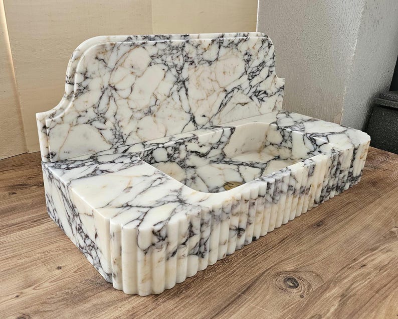 May include: A rectangular marble sink with a white and gray veined pattern. The sink has a curved backsplash and a fluted front. The sink is made of stone and is designed for a bathroom.