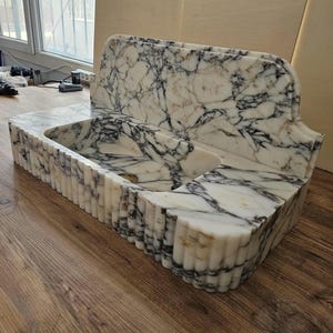 May include: A white marble sink with black veining. The sink has a rectangular basin and a back splash. The front of the sink has a fluted design. The sink is made of natural stone and is a bathroom fixture.