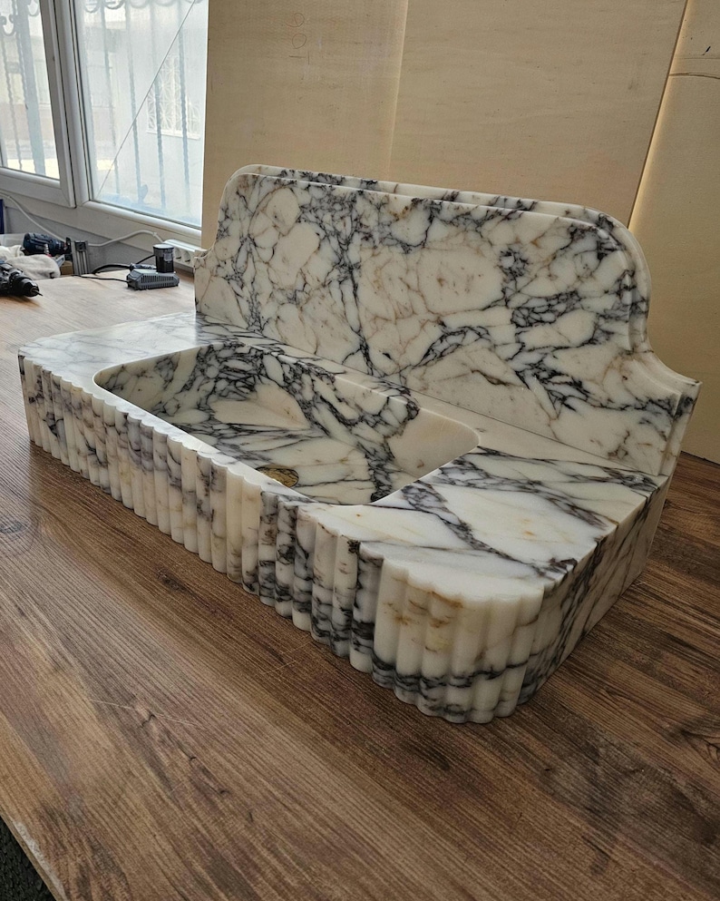 May include: A white marble sink with black veining. The sink has a rectangular basin and a back splash. The front of the sink has a fluted design. The sink is made of natural stone and is a bathroom fixture.
