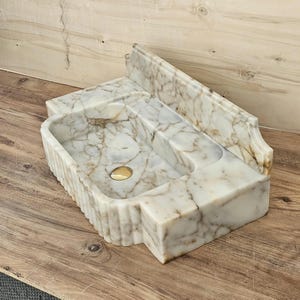 May include: A rectangular marble sink featuring a white base with prominent brown veining. The basin has a smooth interior, a gold-toned drain, and a high back splash. The exterior sides of the sink display a vertical fluted or ribbed texture.