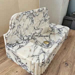 May include: A rectangular marble sink with a white and gray veined pattern. The sink has a built-in backsplash and a fluted front edge. A gold-colored drain is visible in the basin. The sink is made of a polished stone material.