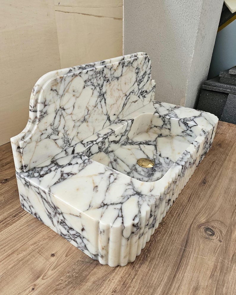 May include: A rectangular marble sink with a white and gray veined pattern. The sink has a built-in backsplash and a fluted front edge. A gold-colored drain is visible in the basin. The sink is made of a polished stone material.