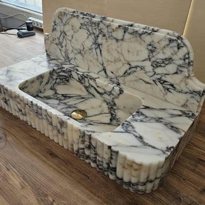 May include: A white marble sink with black veining. The sink has a rectangular basin with a rounded front edge and a decorative back. The front edge of the sink has a fluted design. A gold-colored drain cover is visible.