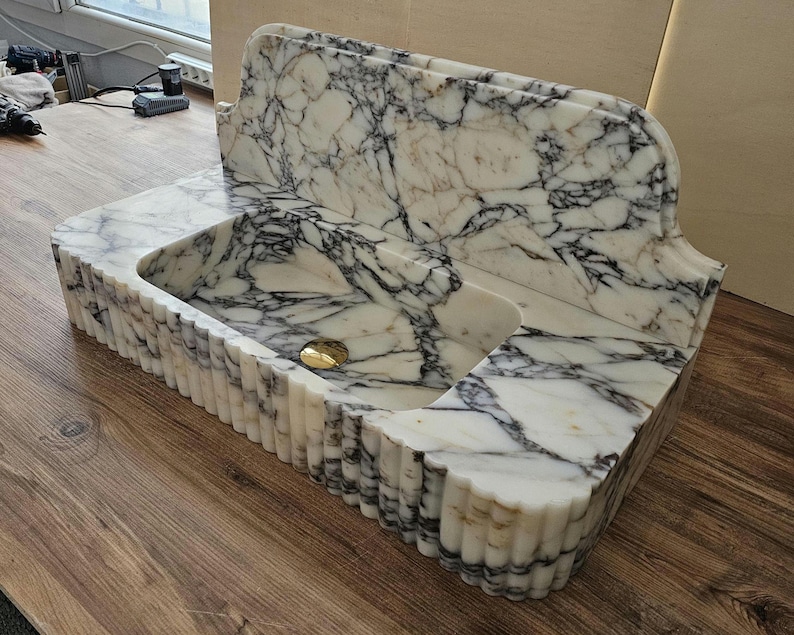 May include: A white marble sink with black veining. The sink has a rectangular basin with a rounded front edge and a decorative back. The front edge of the sink has a fluted design. A gold-colored drain cover is visible.