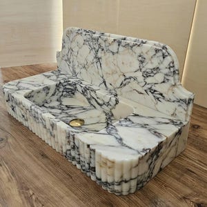 May include: A marble bathroom sink with a white and black veined pattern. The sink has a rectangular basin with a gold-colored drain and a decorative, fluted edge. The back of the sink has a curved, ornate design.