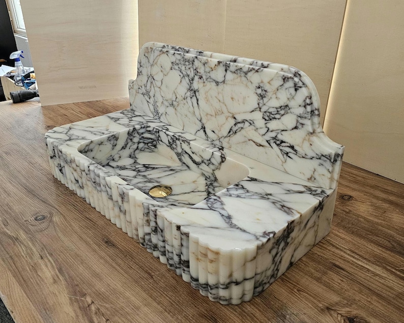 May include: A marble bathroom sink with a white and black veined pattern. The sink has a rectangular basin with a gold-colored drain and a decorative, fluted edge. The back of the sink has a curved, ornate design.