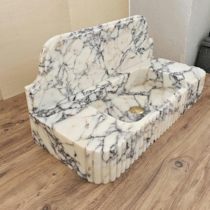 May include: A white marble sink with black veining. The sink has a rectangular basin with a curved back and a fluted front. A gold-colored drain is visible in the basin. The sink is made of stone and is suitable for a bathroom.