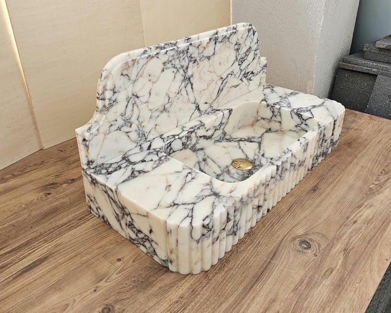 May include: A white marble sink with black veining. The sink has a rectangular basin with a curved back and a fluted front. A gold-colored drain is visible in the basin. The sink is made of stone and is suitable for a bathroom.