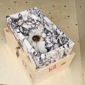 May include: A rectangular marble sink with a black and white pattern sits inside a wooden crate. The sink has a gold-coloured drain cover. The crate is made of light-coloured wood and has a metal latch with a key.