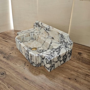 May include: A rectangular marble sink with a white and black veined pattern. The sink has a fluted front and a gold-colored drain. The design includes a back splash and a stepped interior.