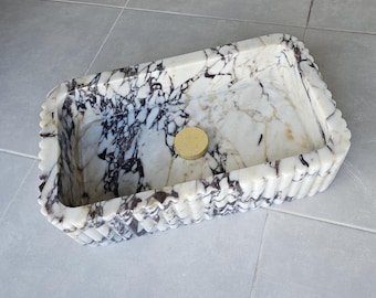 Puede incluir: Un lavabo rectangular de mármol con base blanca y vetas marcadas en color morado oscuro y negro. Las paredes exteriores del recipiente tienen una textura estriada vertical y una tapa de desagüe circular de metal dorado en el centro.